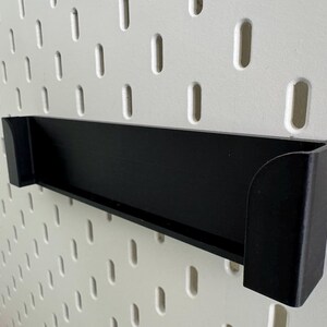 Op de afbeelding: Zwart plastic wandrek met een afgeronde rand. Het rek is gemonteerd op een wit prikbord.