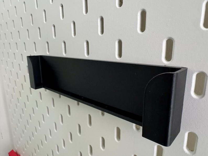 Op de afbeelding: Zwart plastic wandrek met een afgeronde rand. Het rek is gemonteerd op een wit prikbord.