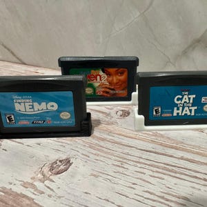 May include: Three Game Boy Advance video game cartridges in black cases, each in a white or black holder. Titles include "Finding Nemo," "Even 2," and "The Cat in the Hat." The cartridges have blue labels with white text.