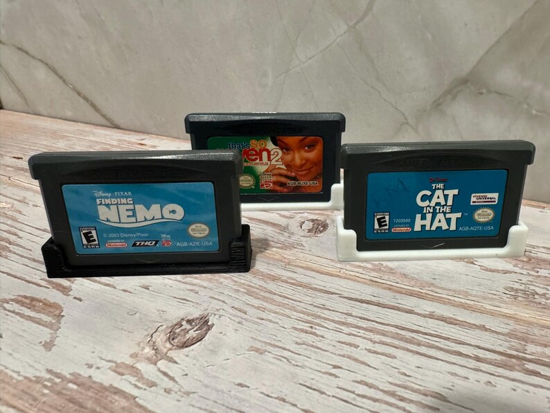 May include: Three Game Boy Advance video game cartridges in black cases, each in a white or black holder. Titles include "Finding Nemo," "Even 2," and "The Cat in the Hat." The cartridges have blue labels with white text.