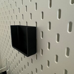 Op de afbeelding: Een zwarte, wandgemonteerde organizer van plastic met twee compartimenten. De organizer is gemonteerd op een wit prikbord.