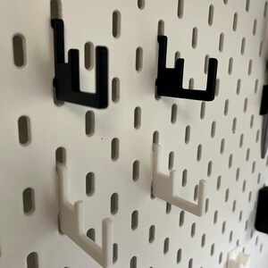 K&ouml;nnte beinhalten: Nahaufnahme einer wei&szlig;en Lochwand mit schwarzen und wei&szlig;en Haken. Die Haken sind so konzipiert, dass sie in die L&ouml;cher der Lochwand passen und eine Aufbewahrungsl&ouml;sung bieten. Die Haken bestehen aus einem robusten Material.
