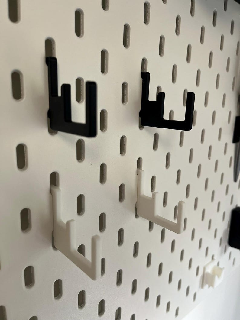 K&ouml;nnte beinhalten: Nahaufnahme einer wei&szlig;en Lochwand mit schwarzen und wei&szlig;en Haken. Die Haken sind so konzipiert, dass sie in die L&ouml;cher der Lochwand passen und eine Aufbewahrungsl&ouml;sung bieten. Die Haken bestehen aus einem robusten Material.