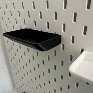 Puede incluir: Dos bandejas de almacenamiento de pl&aacute;stico negro montadas en una pared de tablero de clavijas blanco. Las bandejas son rectangulares y tienen un borde ligeramente curvado.