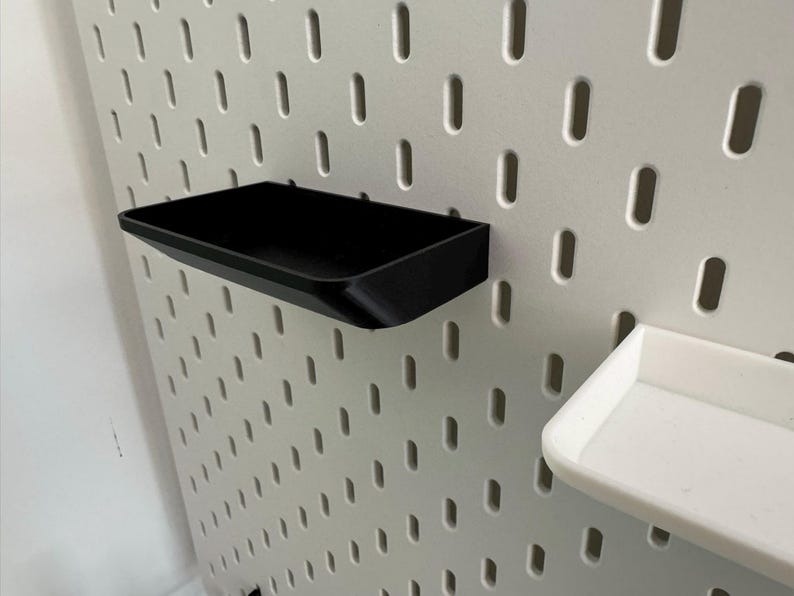 Puede incluir: Dos bandejas de almacenamiento de pl&aacute;stico negro montadas en una pared de tablero de clavijas blanco. Las bandejas son rectangulares y tienen un borde ligeramente curvado.