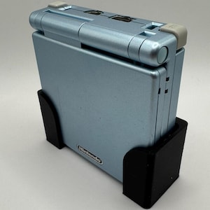 Pu&ograve; includere: Una console per videogiochi portatile Game Boy Advance SP di colore azzurro chiaro con un supporto nero. La console ha un logo Nintendo argentato sul davanti.