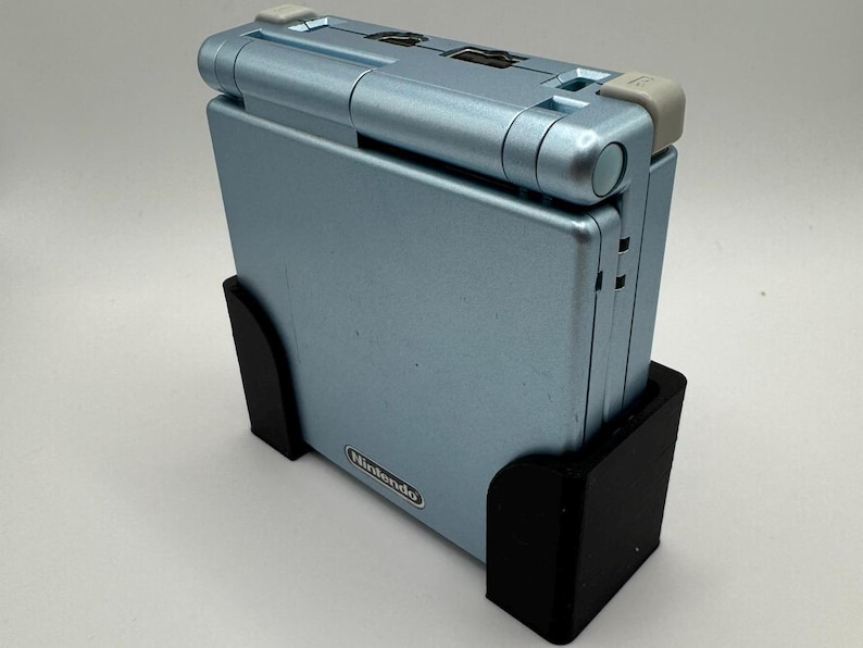 Pu&ograve; includere: Una console per videogiochi portatile Game Boy Advance SP di colore azzurro chiaro con un supporto nero. La console ha un logo Nintendo argentato sul davanti.