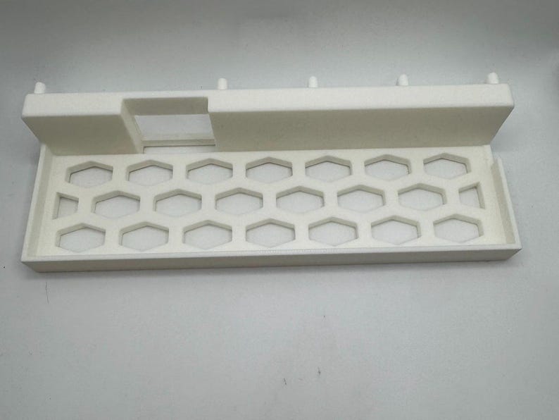Puede incluir: Bandeja blanca rectangular con patr&oacute;n de panal y secci&oacute;n elevada con abertura rectangular. La bandeja tiene peque&ntilde;os pasadores a lo largo del borde superior. Probablemente de pl&aacute;stico.