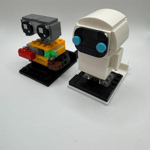 May include: A set of two LEGO minifigures, WALL-E and EVE, from the Disney Pixar movie WALL-E. The figures are posed on black LEGO bases with white accents. WALL-E is yellow, gray, and black with a red toolbox and a green plant. EVE is white with black and blue accents.