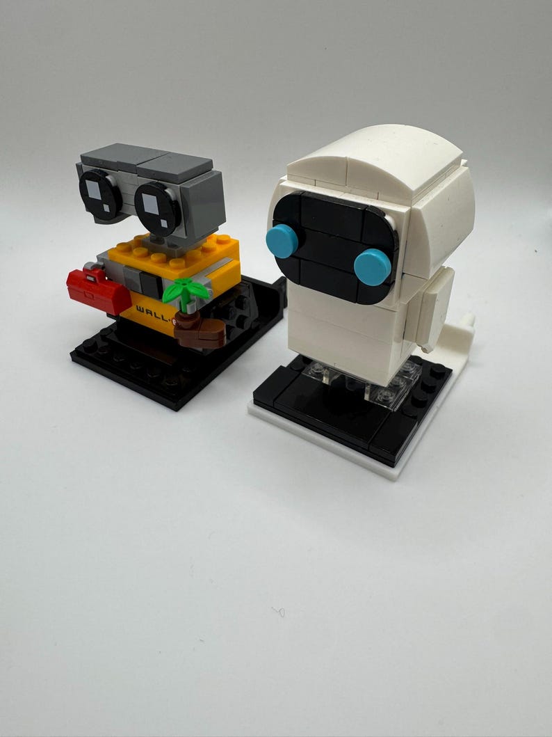 May include: A set of two LEGO minifigures, WALL-E and EVE, from the Disney Pixar movie WALL-E. The figures are posed on black LEGO bases with white accents. WALL-E is yellow, gray, and black with a red toolbox and a green plant. EVE is white with black and blue accents.
