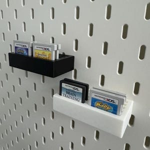 Op de afbeelding: Zwarte en witte wandplanken met Nintendo 3DS-games. De planken tonen games als "Detective Pikachu" en "Brain Training". De achtergrond is een witte pegboard.