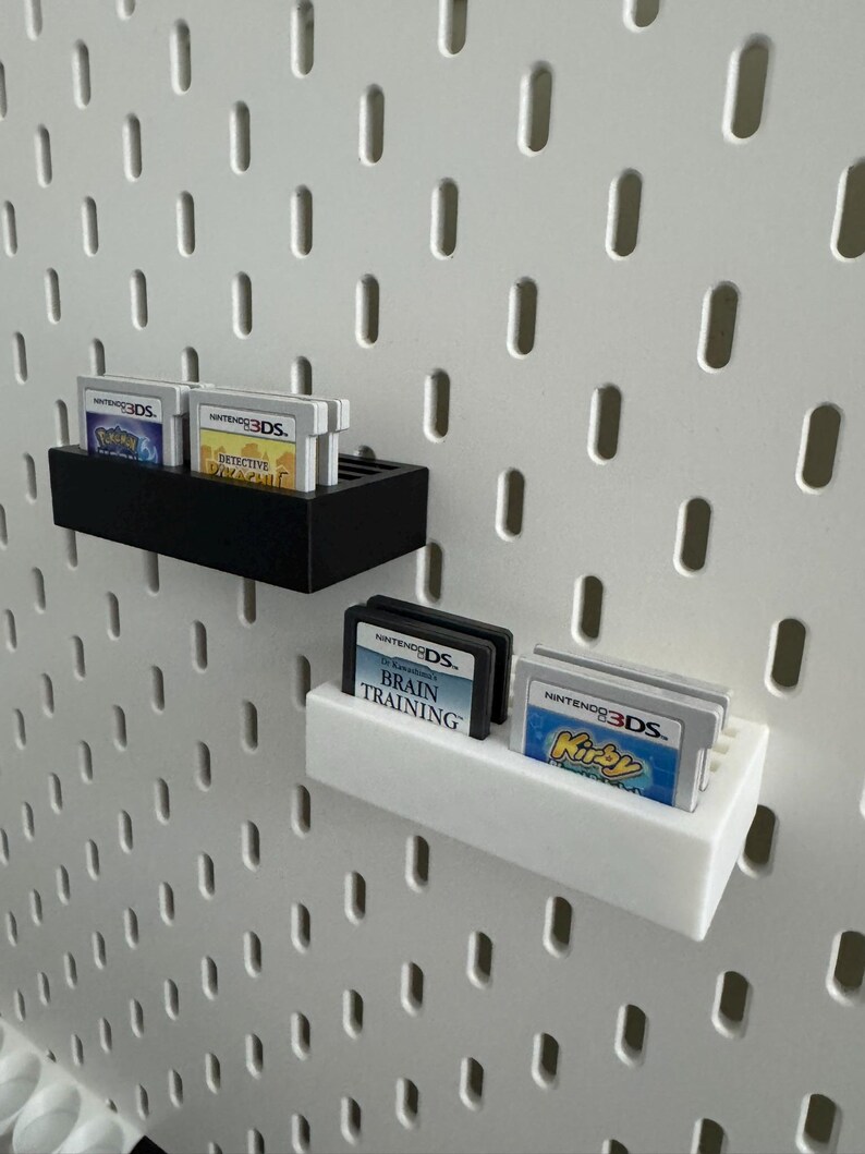 Op de afbeelding: Zwarte en witte wandplanken met Nintendo 3DS-games. De planken tonen games als "Detective Pikachu" en "Brain Training". De achtergrond is een witte pegboard.