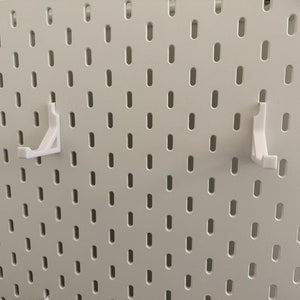 以下が含まれることがあります： 白いペグボードの壁に、白いプラスチック製のペグボードフックが2つ取り付けられています