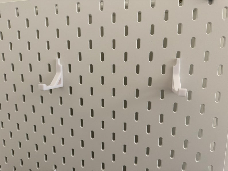 以下が含まれることがあります： 白いペグボードの壁に、白いプラスチック製のペグボードフックが2つ取り付けられています