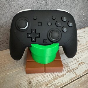 K&ouml;nnte beinhalten: Schwarz-wei&szlig;er Videospiel-Controller auf einem braunen und gr&uuml;nen St&auml;nder. Der Controller hat Kn&ouml;pfe und Joysticks. Der St&auml;nder ist als gr&uuml;ne R&ouml;hre auf einem braunen Block gestaltet.