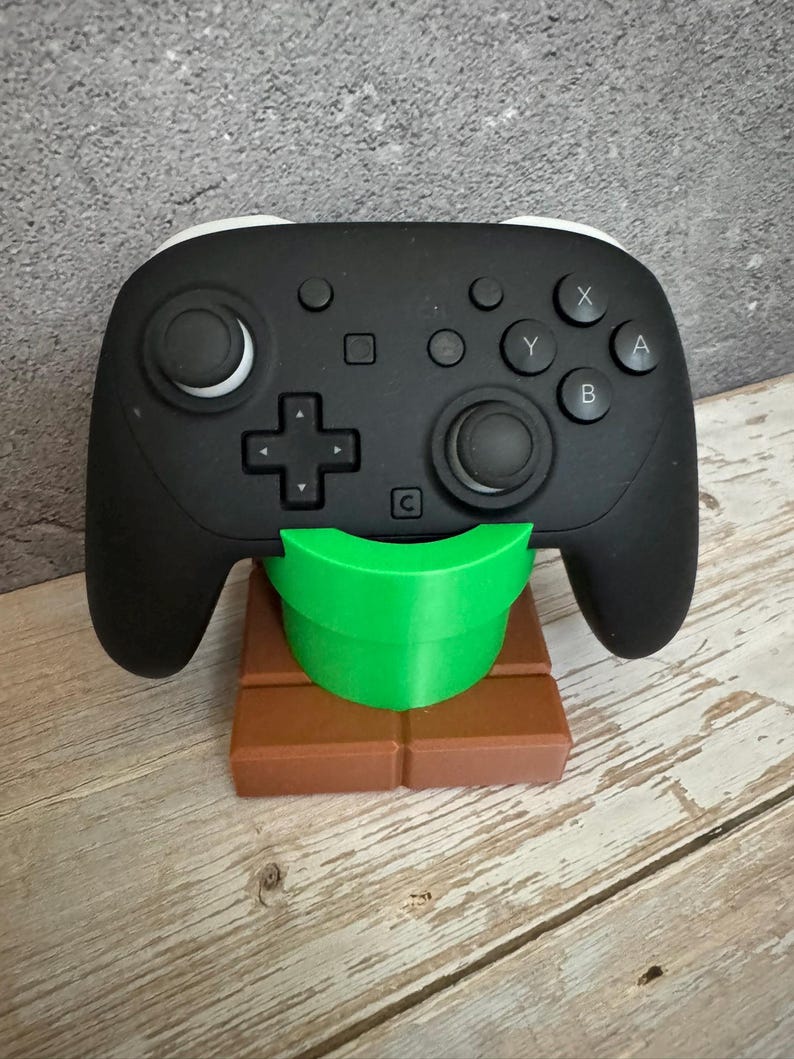 K&ouml;nnte beinhalten: Schwarz-wei&szlig;er Videospiel-Controller auf einem braunen und gr&uuml;nen St&auml;nder. Der Controller hat Kn&ouml;pfe und Joysticks. Der St&auml;nder ist als gr&uuml;ne R&ouml;hre auf einem braunen Block gestaltet.