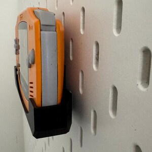 Op de afbeelding: Een zwarte wandhouder met een oranje en grijze handheld-videogames console bevestigd. De console heeft knoppen en een scherm.