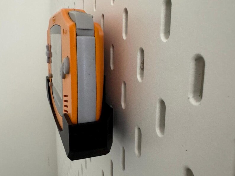 Op de afbeelding: Een zwarte wandhouder met een oranje en grijze handheld-videogames console bevestigd. De console heeft knoppen en een scherm.