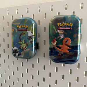 K&ouml;nnte beinhalten: Zwei Pok&eacute;mon Trading Card Game-Dosen. Die Dosen sind rechteckig mit abgerundeten Kanten und zeigen farbenfrohe Illustrationen von Pok&eacute;mon-Charakteren. Die Dosen sind an einer wei&szlig;en Lochwand befestigt. Das Pok&eacute;mon-Logo und der Text "Trading Card Game" sind sichtbar.