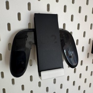 Op de afbeelding: Zwarte gamecontrollerhouder met een witte basis, gemonteerd op een wit pegboard. De houder is ontworpen om een gamecontroller en een mobiel apparaat veilig vast te houden. Het pegboard heeft gelijkmatig verdeelde ovale gaten.