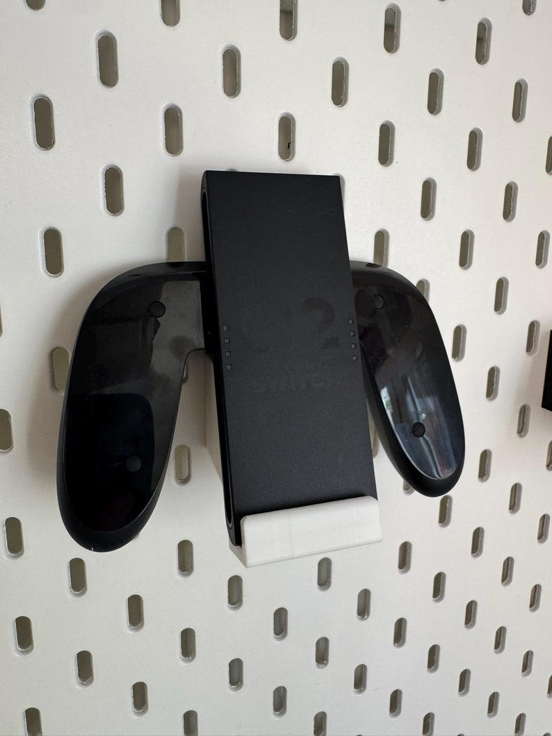 Op de afbeelding: Zwarte gamecontrollerhouder met een witte basis, gemonteerd op een wit pegboard. De houder is ontworpen om een gamecontroller en een mobiel apparaat veilig vast te houden. Het pegboard heeft gelijkmatig verdeelde ovale gaten.