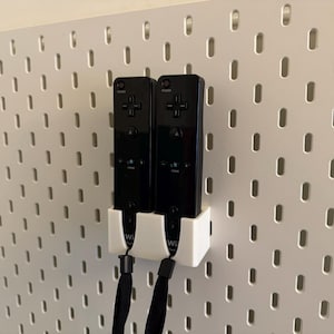 Puede incluir: Dos mandos de Wii negros est&aacute;n montados en una pared perforada blanca con un soporte blanco. Los mandos tienen correas de mu&ntilde;eca negras y est&aacute;n colocados verticalmente. La pared perforada tiene agujeros espaciados.
