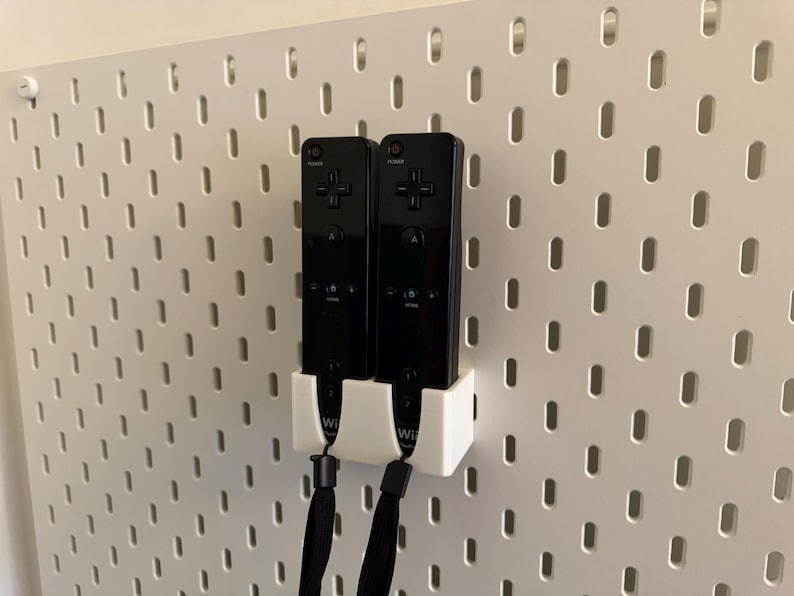 Puede incluir: Dos mandos de Wii negros est&aacute;n montados en una pared perforada blanca con un soporte blanco. Los mandos tienen correas de mu&ntilde;eca negras y est&aacute;n colocados verticalmente. La pared perforada tiene agujeros espaciados.