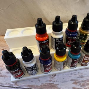Puede incluir: Un soporte de pl&aacute;stico blanco para botes de pintura con diez botes de pintura peque&ntilde;os. Los botes son de varios colores, como rojo, blanco, azul y amarillo. Los botes est&aacute;n etiquetados con la marca "Vallejo" y "Game Color".