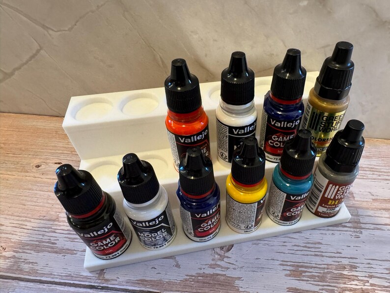 Puede incluir: Un soporte de pl&aacute;stico blanco para botes de pintura con diez botes de pintura peque&ntilde;os. Los botes son de varios colores, como rojo, blanco, azul y amarillo. Los botes est&aacute;n etiquetados con la marca "Vallejo" y "Game Color".