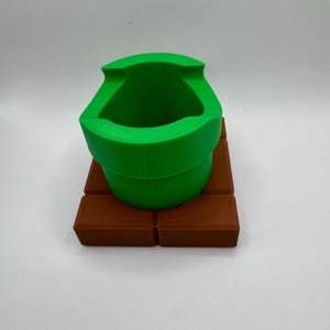 K&ouml;nnte beinhalten: Ein gr&uuml;ner Rohrtopf, der an ein Videospiel erinnert, steht auf einer braunen, ziegelartigen Basis. Der Topf hat eine breite &Ouml;ffnung und ein gebogenes Design. Die Basis besteht aus mehreren rechteckigen Bl&ouml;cken.