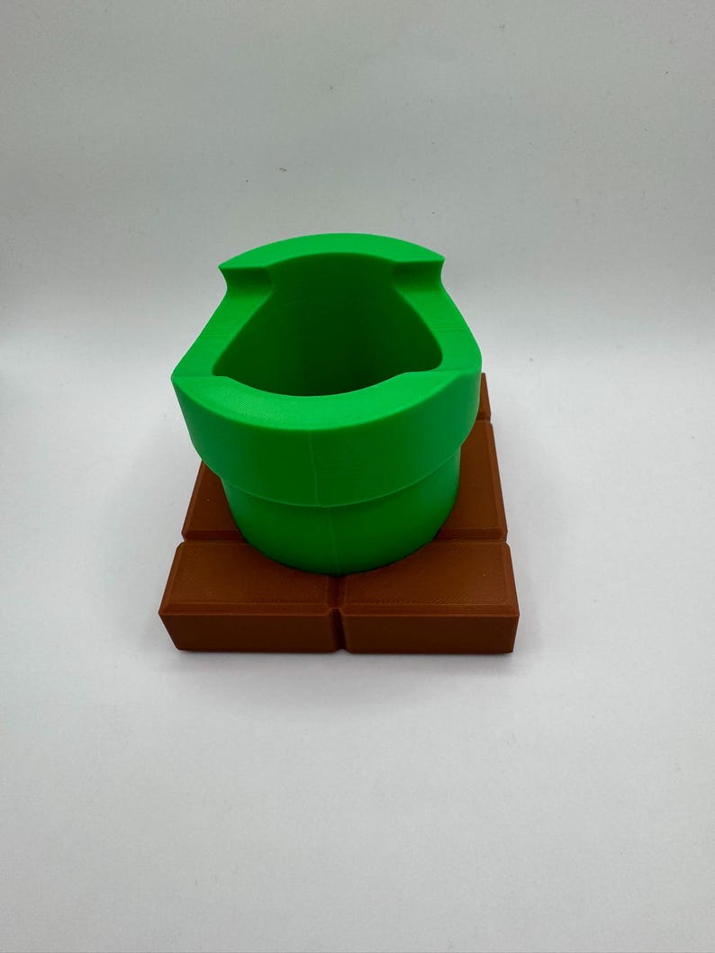 K&ouml;nnte beinhalten: Ein gr&uuml;ner Rohrtopf, der an ein Videospiel erinnert, steht auf einer braunen, ziegelartigen Basis. Der Topf hat eine breite &Ouml;ffnung und ein gebogenes Design. Die Basis besteht aus mehreren rechteckigen Bl&ouml;cken.