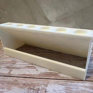 Puede incluir: Un soporte blanco rectangular con m&uacute;ltiples aberturas circulares, posiblemente para bol&iacute;grafos u objetos peque&ntilde;os. El soporte est&aacute; hecho de un material s&oacute;lido y opaco y se encuentra sobre una superficie de madera. El dise&ntilde;o presenta un lado inclinado y una base rectangular.