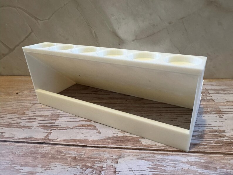 Puede incluir: Un soporte blanco rectangular con m&uacute;ltiples aberturas circulares, posiblemente para bol&iacute;grafos u objetos peque&ntilde;os. El soporte est&aacute; hecho de un material s&oacute;lido y opaco y se encuentra sobre una superficie de madera. El dise&ntilde;o presenta un lado inclinado y una base rectangular.