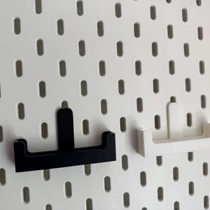 Op de afbeelding: Twee pegboardplanken, &eacute;&eacute;n zwart en &eacute;&eacute;n wit, worden getoond tegen een witte pegboardachtergrond. De planken hebben een eenvoudig, modern ontwerp, perfect voor het organiseren van kleine items. Het pegboard heeft gelijkmatig verdeelde ovale gaten.