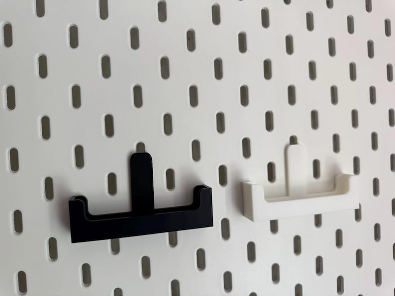 Op de afbeelding: Twee pegboardplanken, &eacute;&eacute;n zwart en &eacute;&eacute;n wit, worden getoond tegen een witte pegboardachtergrond. De planken hebben een eenvoudig, modern ontwerp, perfect voor het organiseren van kleine items. Het pegboard heeft gelijkmatig verdeelde ovale gaten.