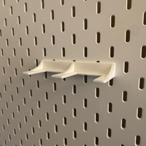 May include: Three white pegboard shelf brackets are mounted on a white pegboard. The brackets have a simple, angular design with a small lip to hold items. The pegboard has a grid of evenly spaced holes.
