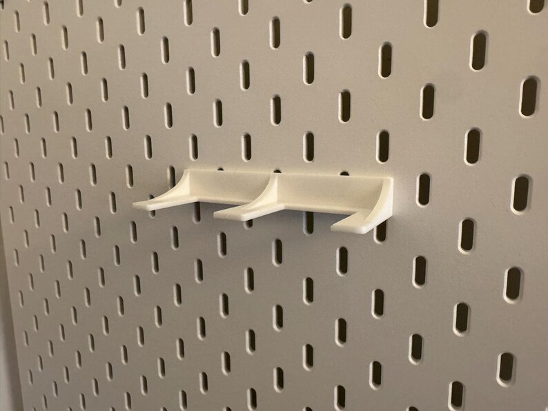 May include: Three white pegboard shelf brackets are mounted on a white pegboard. The brackets have a simple, angular design with a small lip to hold items. The pegboard has a grid of evenly spaced holes.