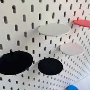 Op de afbeelding: Vijf ronde planken in verschillende kleuren gemonteerd op een witte prikbordwand. De planken zijn zwart, wit en roze.