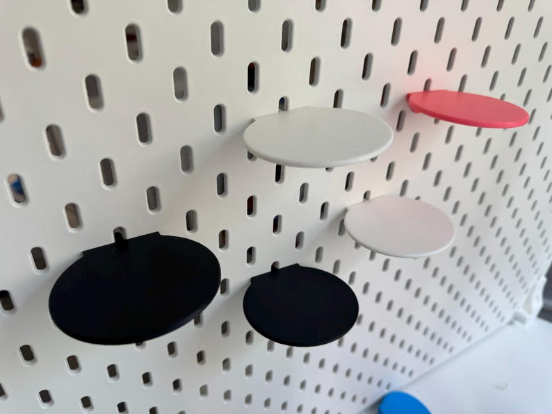 Op de afbeelding: Vijf ronde planken in verschillende kleuren gemonteerd op een witte prikbordwand. De planken zijn zwart, wit en roze.