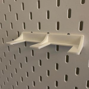 May include: A white pegboard shelf bracket, designed to attach to a pegboard. The bracket has a simple, functional design with two support arms. The pegboard is white with evenly spaced oval holes.