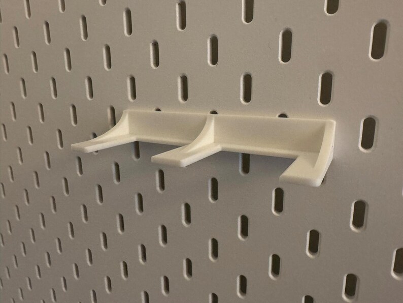 May include: A white pegboard shelf bracket, designed to attach to a pegboard. The bracket has a simple, functional design with two support arms. The pegboard is white with evenly spaced oval holes.