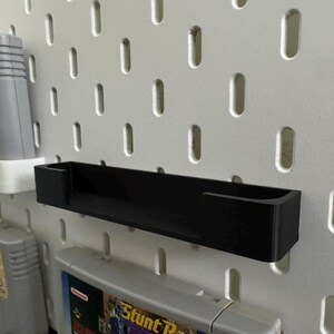 May include: A black, rectangular shelf for video game cartridges, mounted on a white pegboard. Several game cartridges are visible. The shelf has a simple, minimalist design, suitable for organizing retro games.
