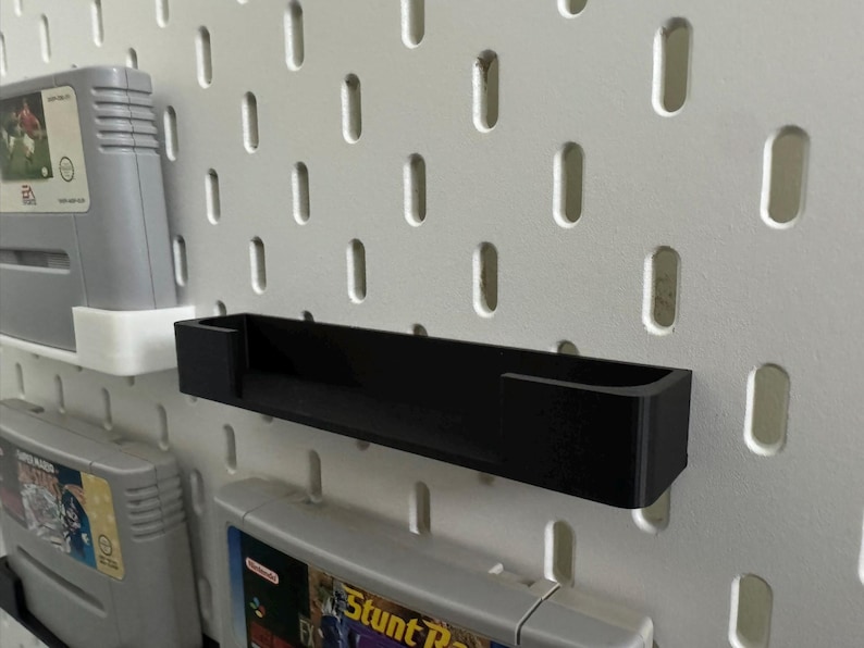 May include: A black, rectangular shelf for video game cartridges, mounted on a white pegboard. Several game cartridges are visible. The shelf has a simple, minimalist design, suitable for organizing retro games.