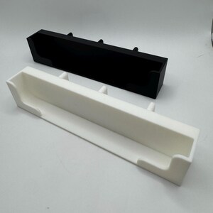 May include: Two rectangular trays, one black and one white, are displayed. Each tray has a trough-like design with raised supports. The white tray is positioned in front of the black tray. The trays are made of a solid material and are set against a plain background.