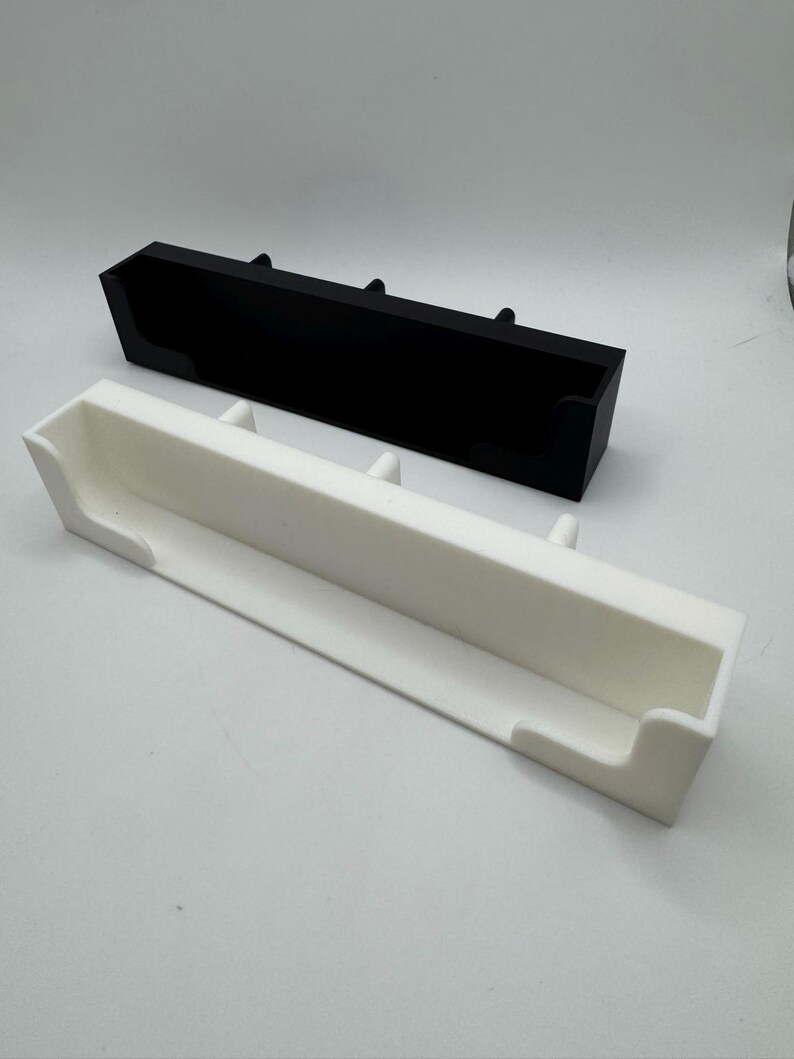 May include: Two rectangular trays, one black and one white, are displayed. Each tray has a trough-like design with raised supports. The white tray is positioned in front of the black tray. The trays are made of a solid material and are set against a plain background.
