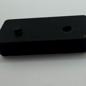 May include: A black plastic rectangular block with two holes on the top. The block has a textured surface.