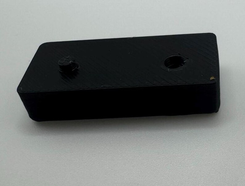 May include: A black plastic rectangular block with two holes on the top. The block has a textured surface.