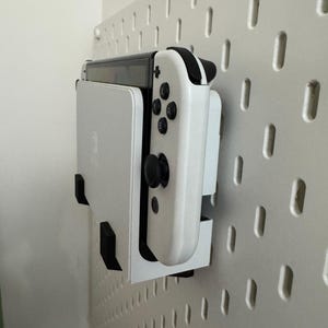 May include: A white Nintendo Switch console and controller are mounted on a white pegboard. The controller has black buttons and joysticks. The pegboard has a series of evenly spaced, vertical slots.