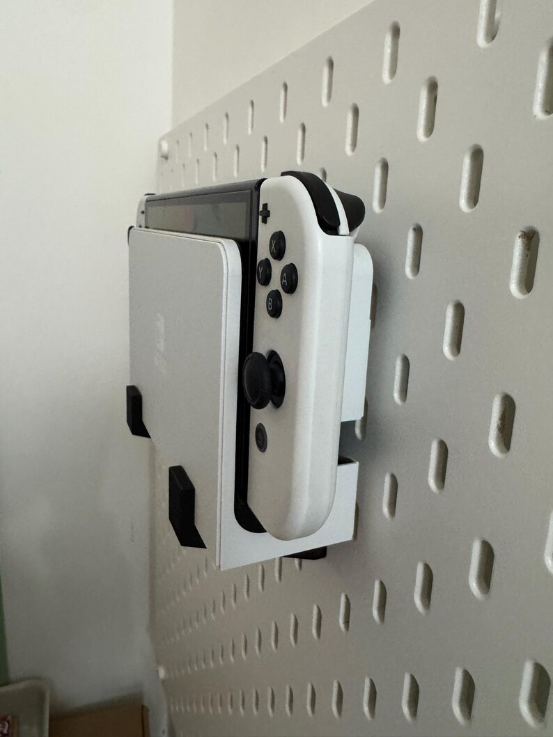 May include: A white Nintendo Switch console and controller are mounted on a white pegboard. The controller has black buttons and joysticks. The pegboard has a series of evenly spaced, vertical slots.