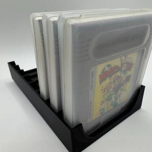 K&ouml;nnte beinhalten: Ein schwarzer Game-Cartridge-Halter aus Kunststoff mit vier transparenten Game-Cartridges aus Kunststoff. Die Game-Cartridges haben ein gelbes und farbenfrohes Etikett. Der Halter ist f&uuml;r mehrere Game-Cartridges konzipiert.